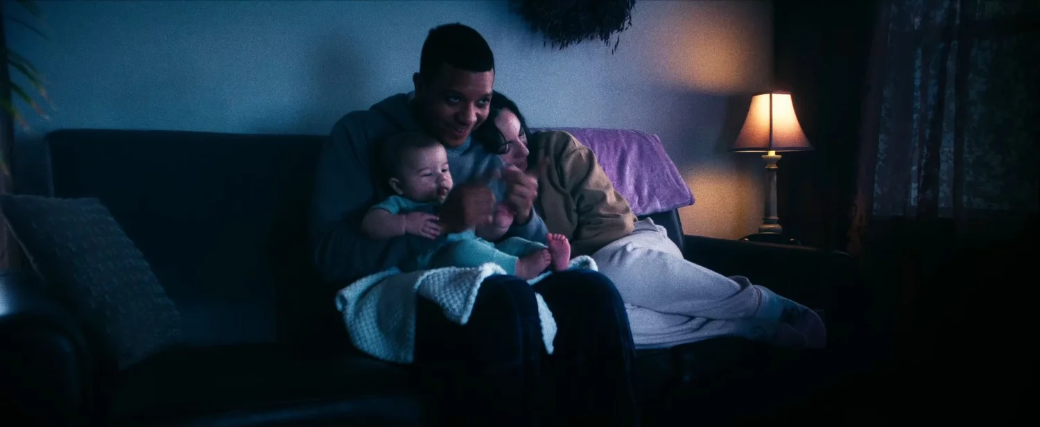 Still from INERTIA: a man on a dark couch holds a baby on his lap while a woman curls up against his shoulder, a table lamp glowing beside them.