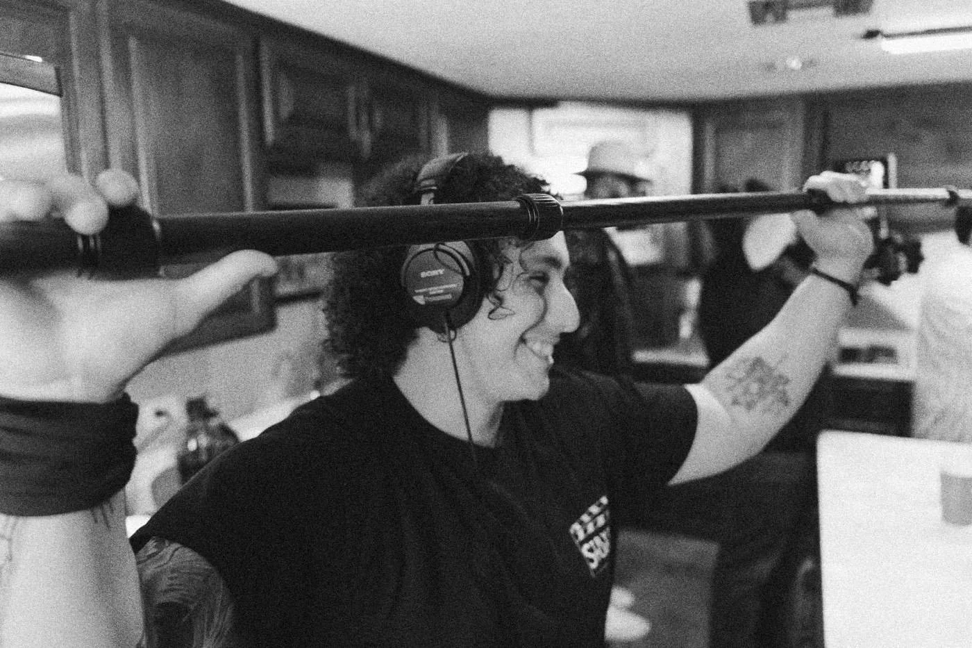 Behind the scenes on INERTIA: a boom operator in red light raises the pole overhead and smiles.
