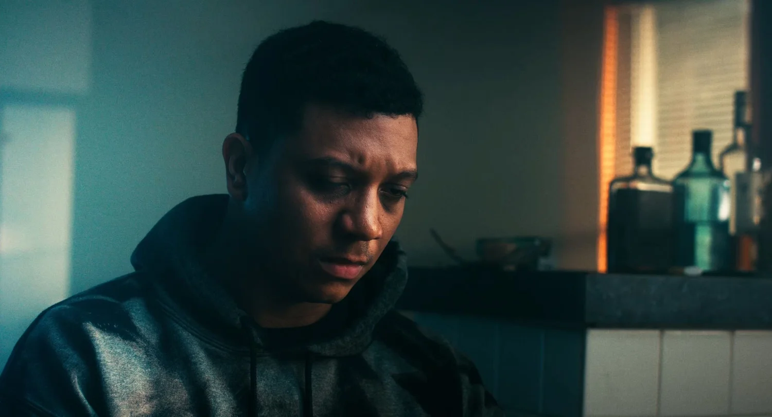 Still from INERTIA: a man in a dark hooded sweatshirt looks down in a dim kitchen, glass bottles lined up on the counter behind him.