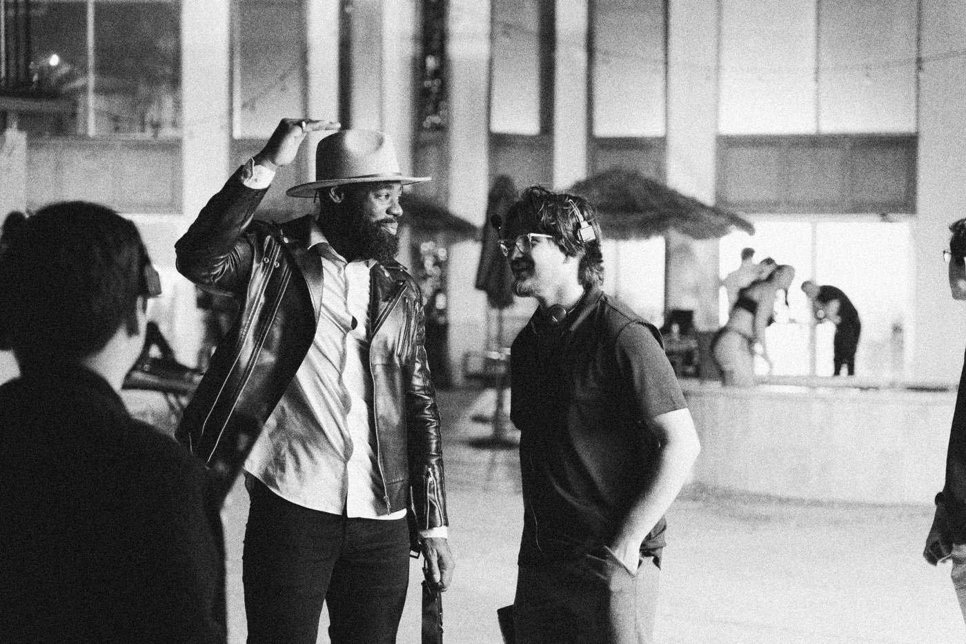 Behind the scenes on INERTIA: an actor in a hat and leather jacket laughs with crew members on a patio in daylight.