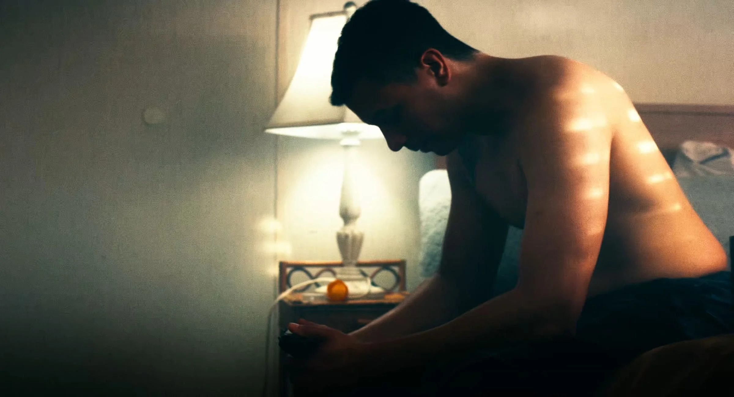 Still from INERTIA: a shirtless man sits on the edge of a bed with his head bowed beside a lit lamp on a nightstand.