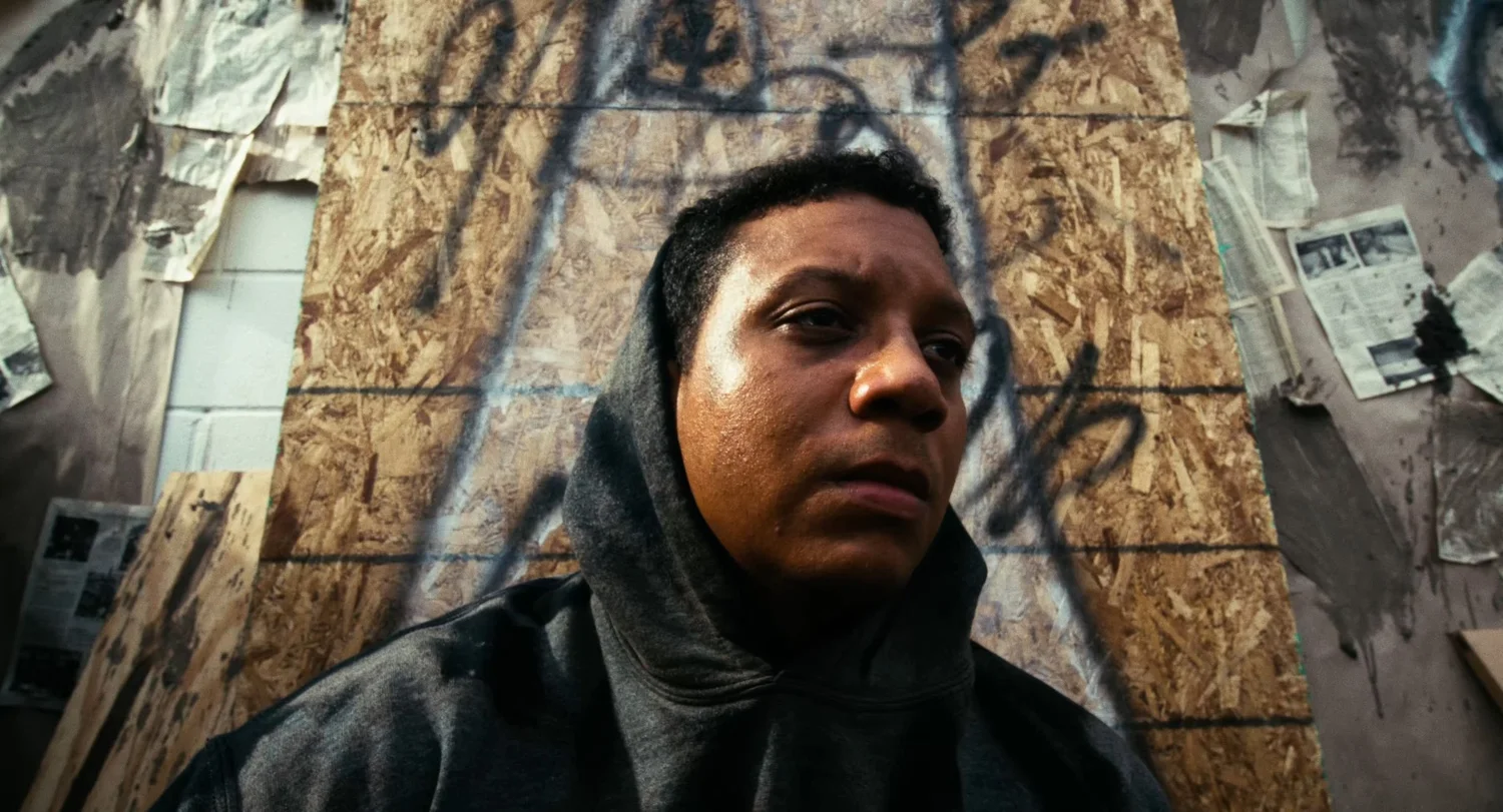Still from INERTIA: a close view of a person in a gray hooded sweatshirt in front of a plywood board with graffiti, looking off to the right.