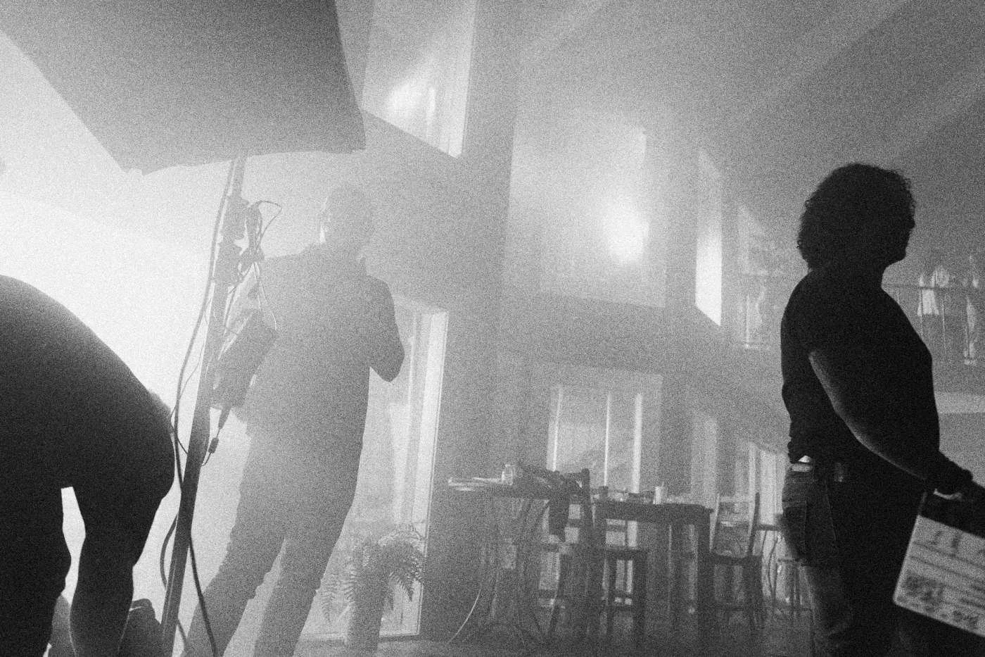 Behind the scenes on INERTIA: crew members stand in silhouette under a large light in a hazy two level room.