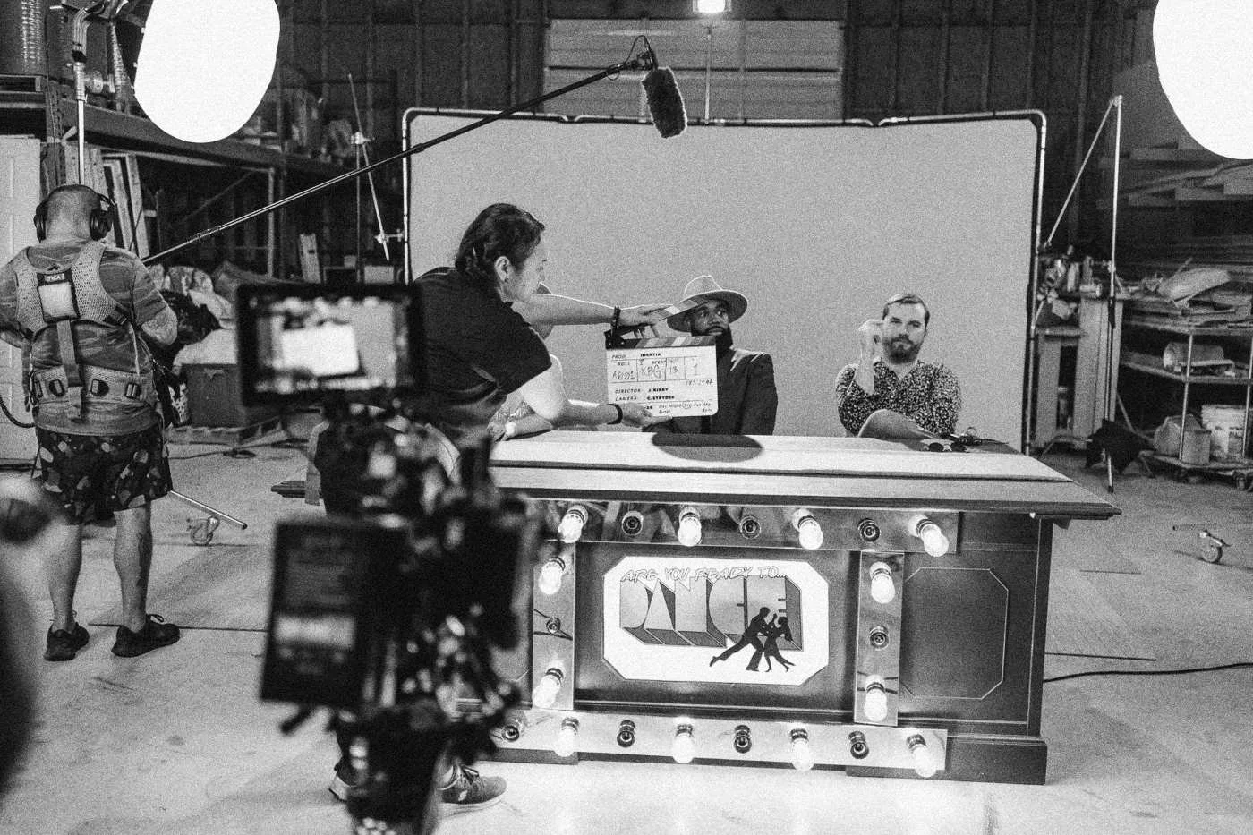 Behind the scenes on INERTIA: a slate is held up at a lit game show desk on a green screen stage while the camera rolls.