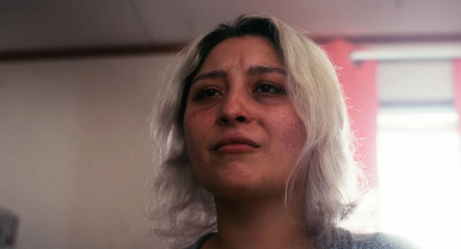 Still from INERTIA: a low angle close view of a woman with platinum blonde hair and freckles looking past the camera, a red curtain and bright window behind her.