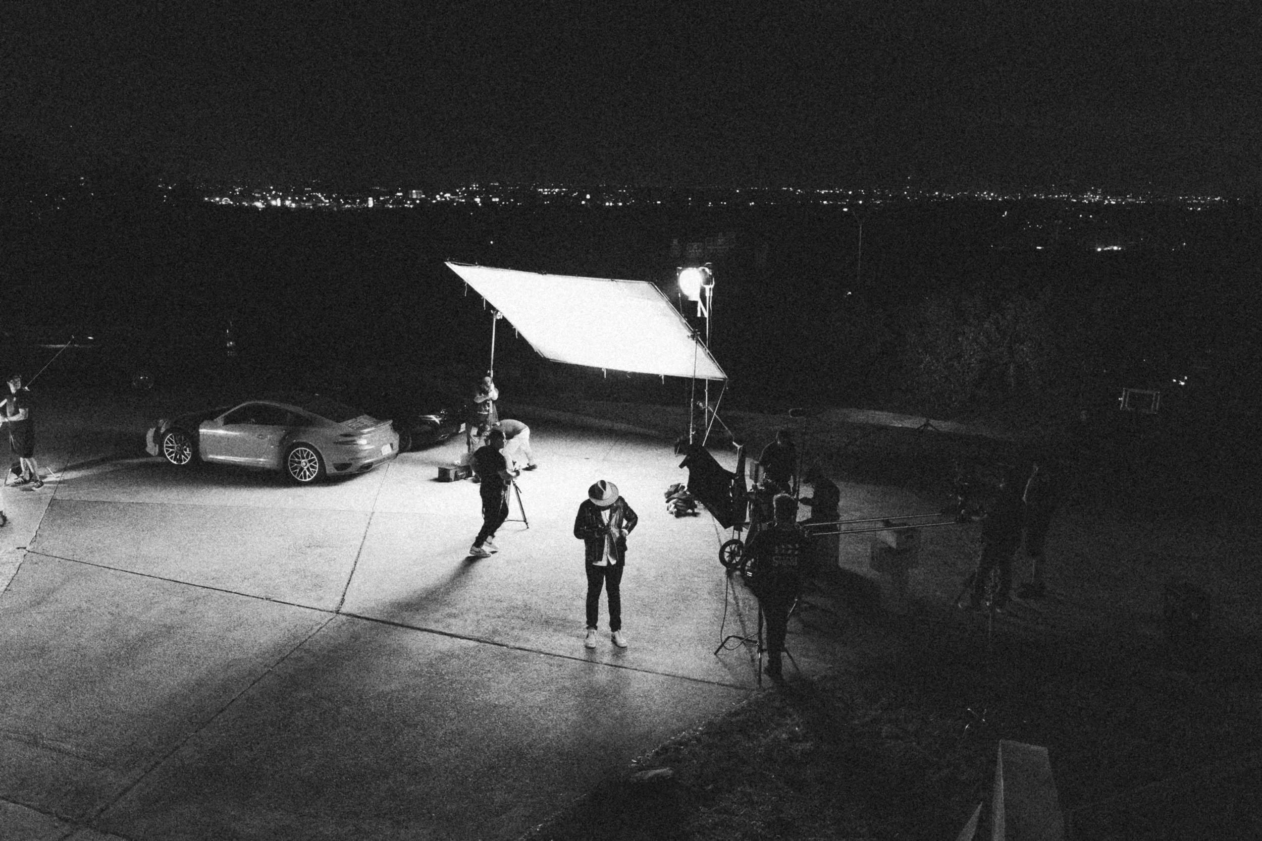 Behind the scenes on INERTIA: seen from above at night, a man in a hat stands in a pool of light on a driveway beneath a large overhead silk while crew work around him, sports cars parked behind and city lights along the horizon.