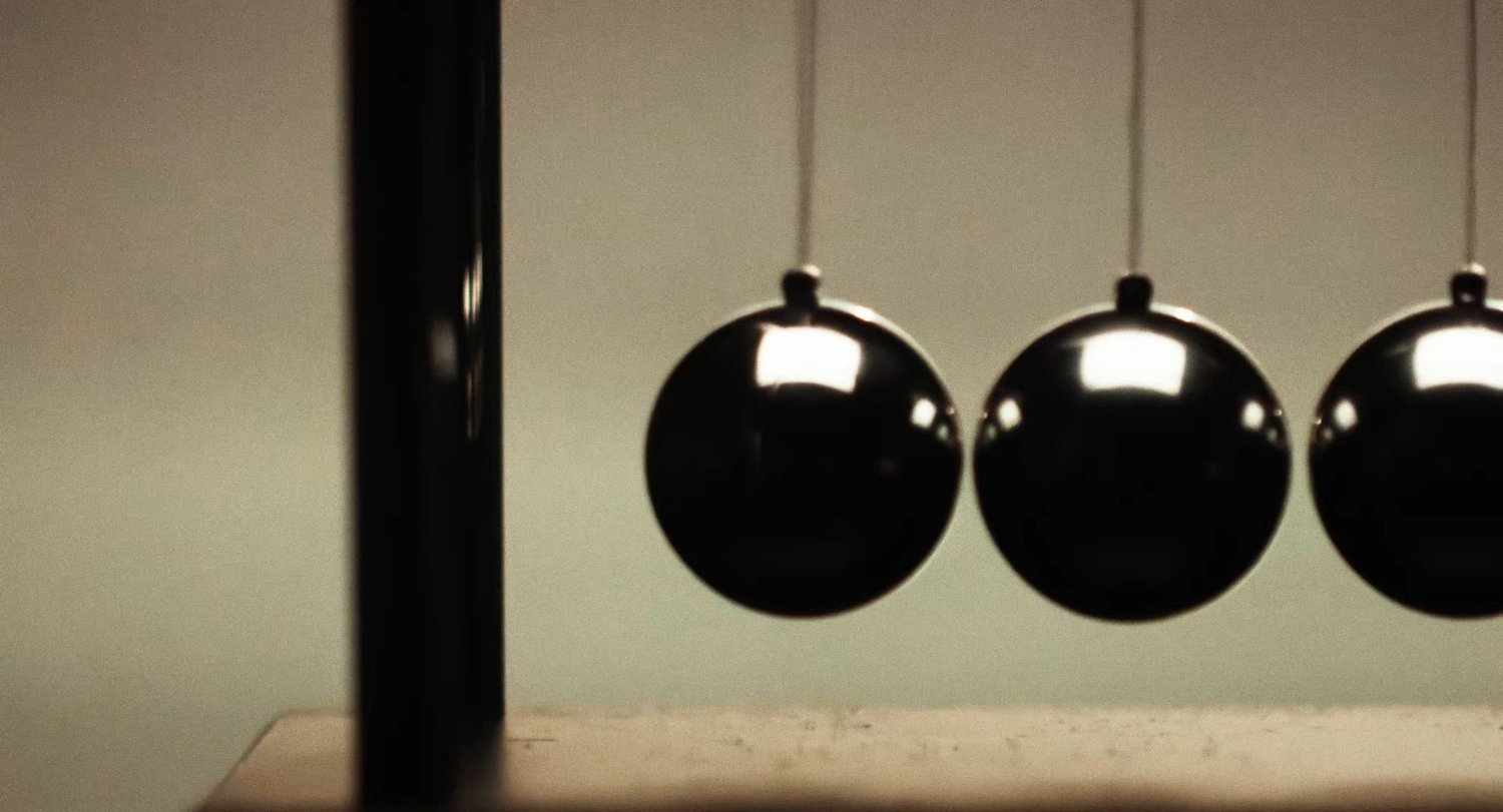 Still from INERTIA: a Newton's cradle, three polished metal balls hanging from thin strings beside its dark upright post, against a pale wall.