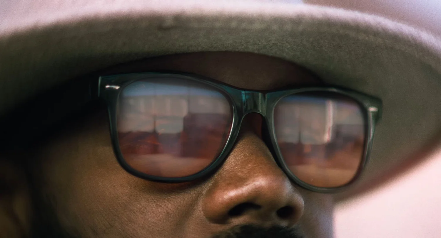 Still from INERTIA: an extreme close view of a man in a brimmed hat and dark sunglasses that reflect a street.