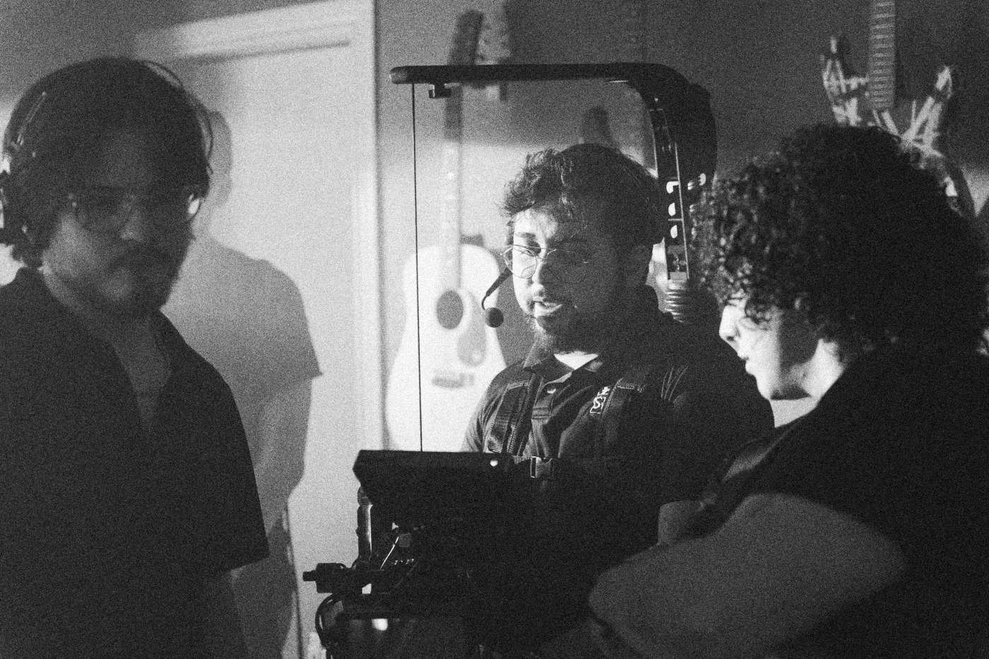 Behind the scenes on INERTIA: three crew members in headsets confer beside a camera rig, guitars on the wall behind.