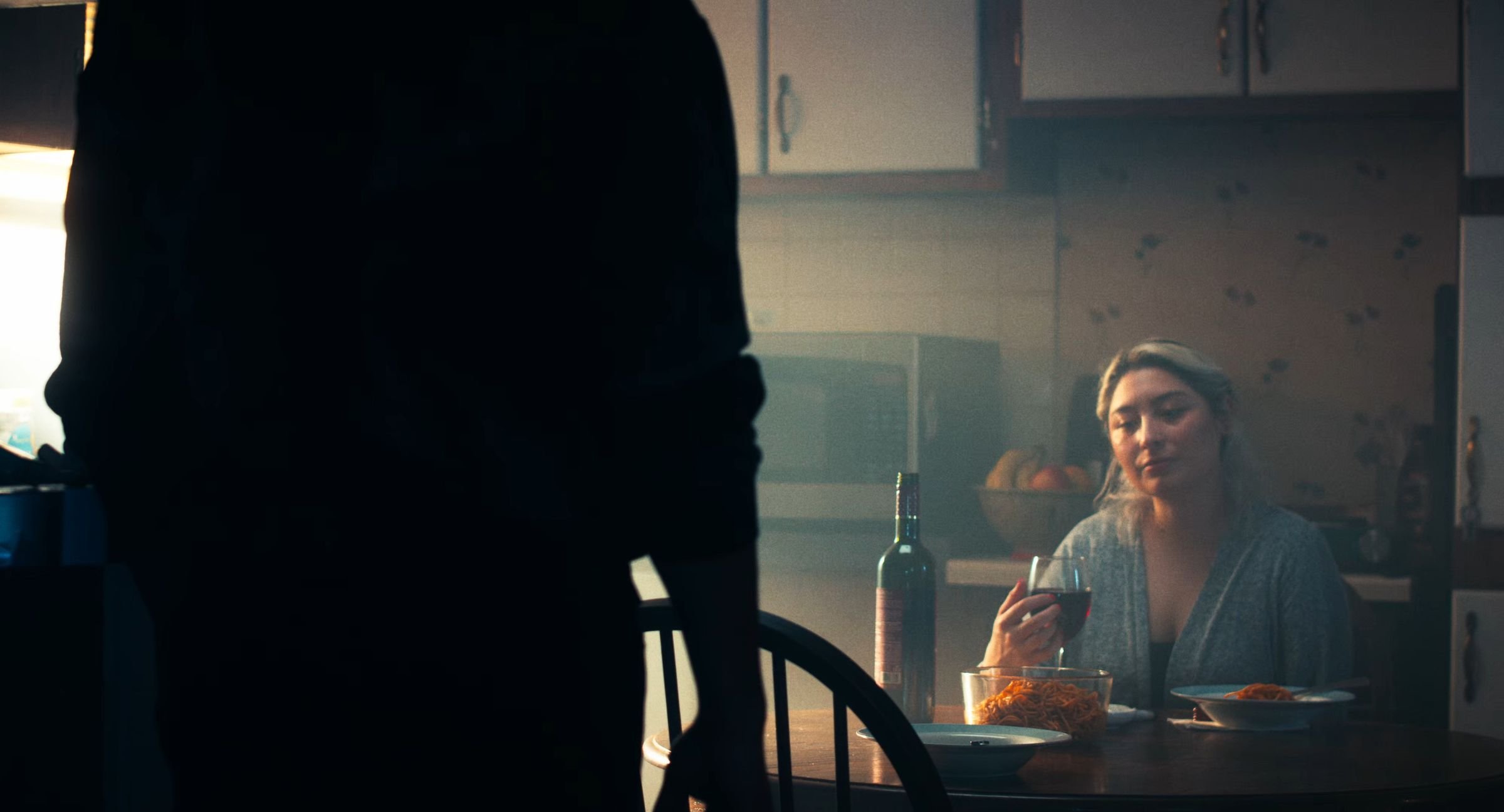 Still from INERTIA: a woman at a kitchen table holds a glass of red wine beside a wine bottle and bowls of spaghetti while a dark figure stands in the foreground.