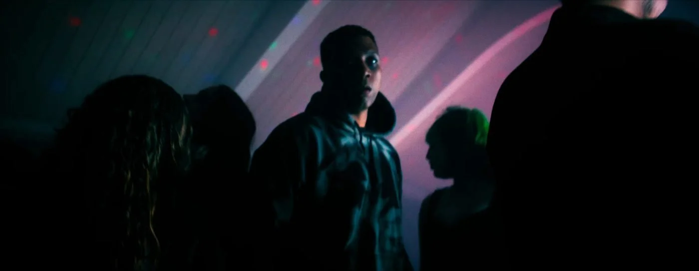 Still from INERTIA: a man in a dark hooded sweatshirt stands among silhouetted people under pink and purple party lights.