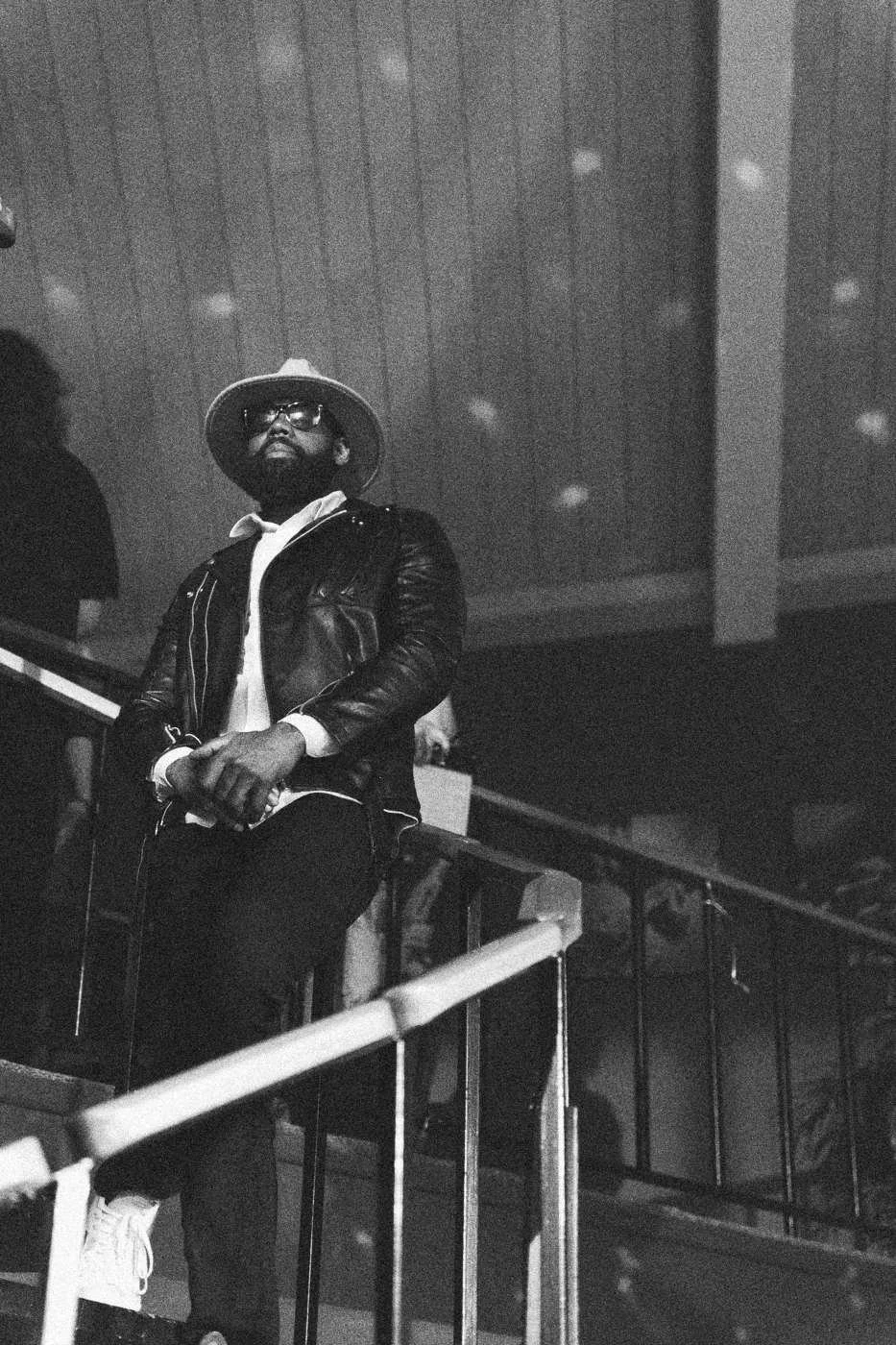 Behind the scenes on INERTIA: an actor in a hat and leather jacket on a staircase under colored party lights.