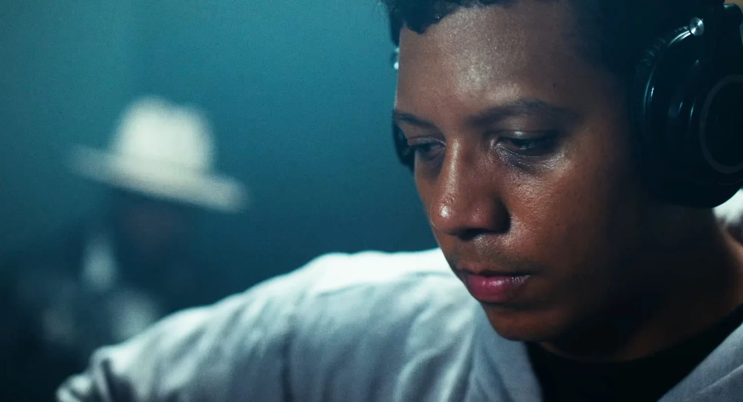 Still from INERTIA: a close view of a man wearing headphones looking down, with a blurred figure in a white hat behind him.