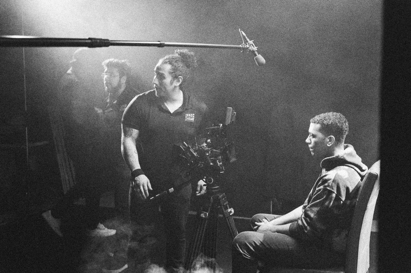 Behind the scenes on INERTIA: a camera operator and boom work close to an actor seated in a red chair in light haze.
