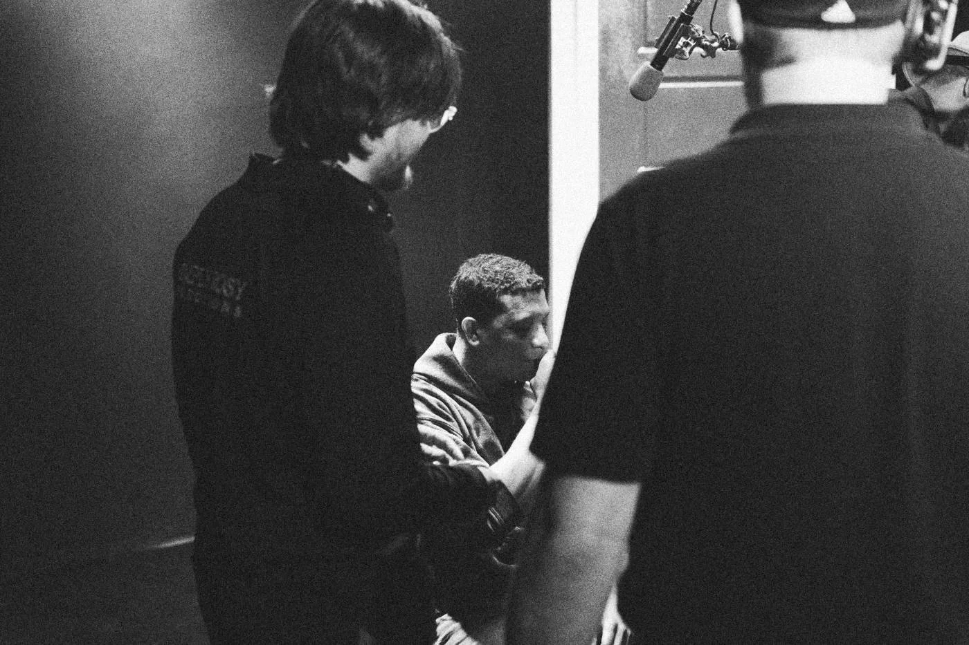 Behind the scenes on INERTIA: two crew members talk with a seated actor beside a red door, one seen from behind.