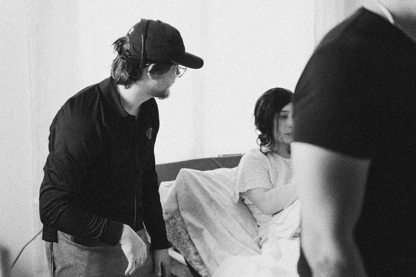 Behind the scenes on INERTIA: a crew member in a cap leans in to talk with an actress lying in a bed on set.