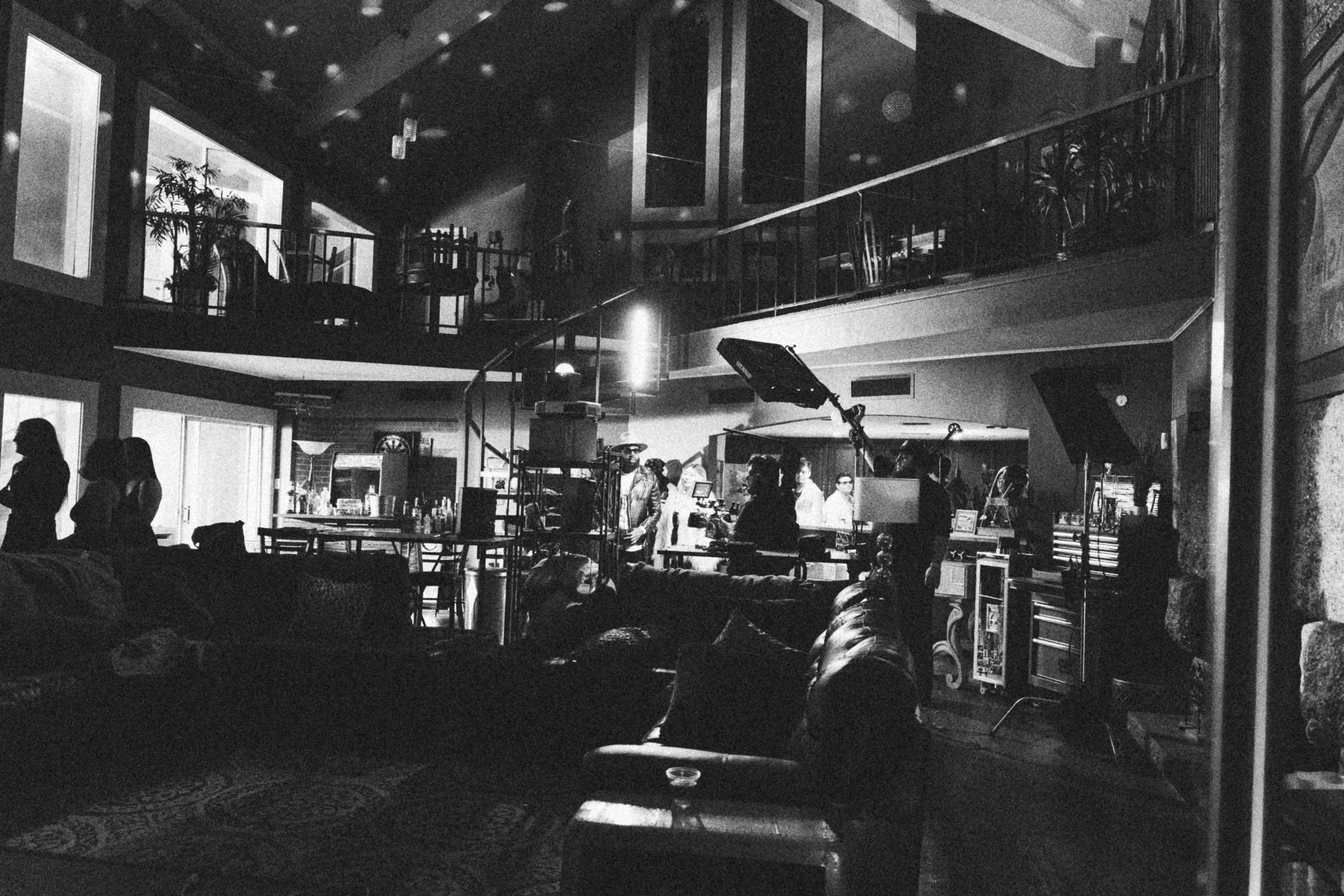 Behind the scenes on INERTIA: a large two story living room at night, dark couches in front and crew with a camera, a softbox and lights working at the far end beneath a balcony.