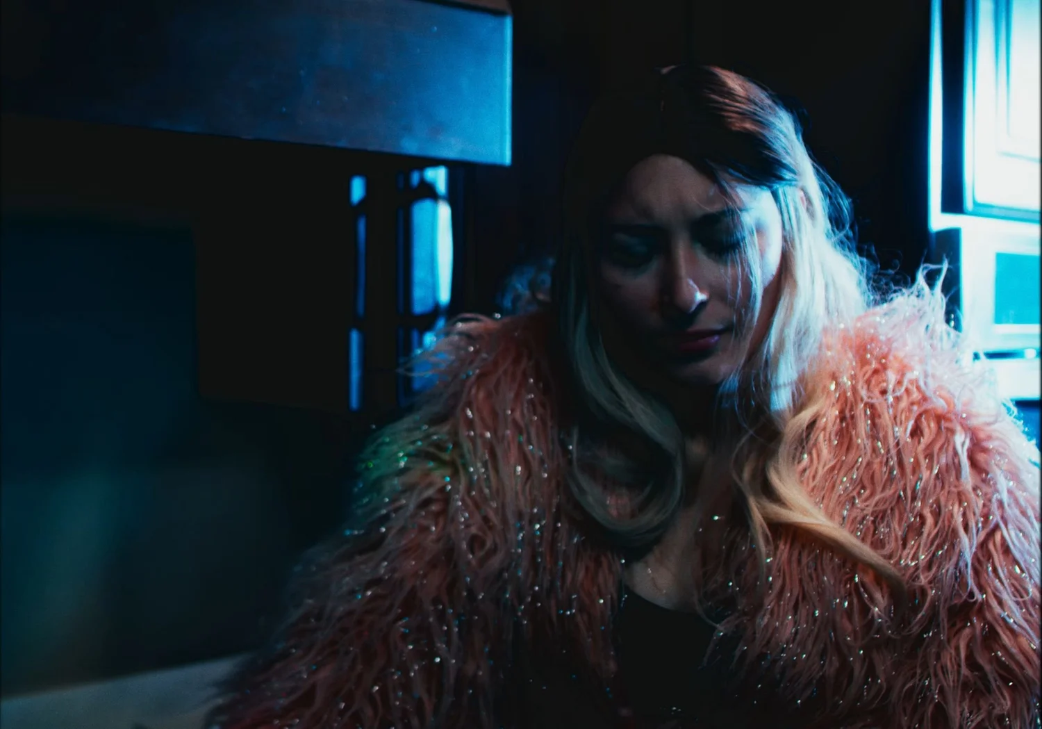 Still from INERTIA: a woman with long two tone hair in a pink shaggy sparkling coat looks down, lit by blue light from a window.