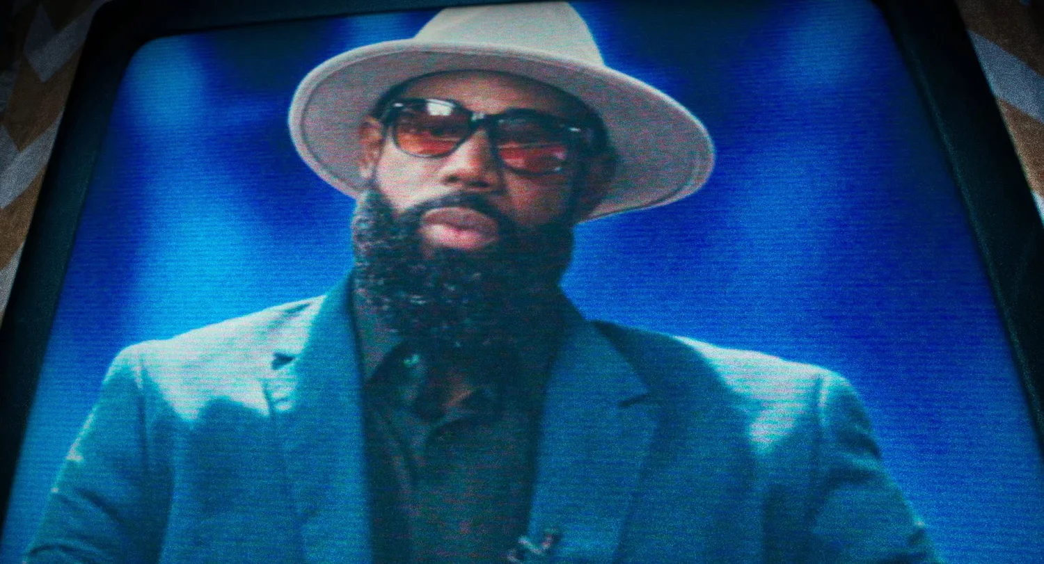 Still from INERTIA: a close view of a television screen showing a bearded man in a light hat, tinted sunglasses and a teal jacket.