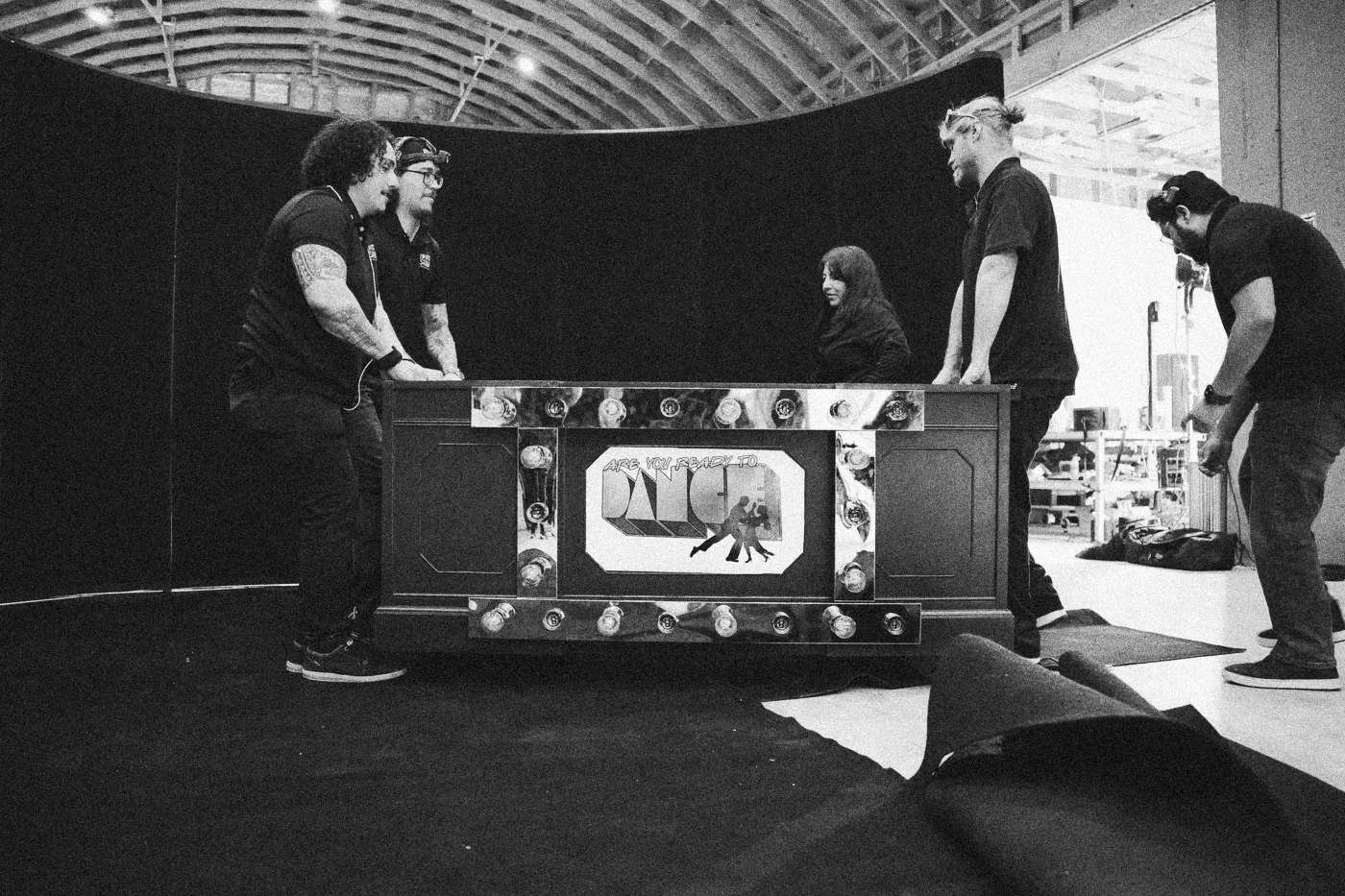 Behind the scenes on INERTIA: crew members move a decorated set piece across a black draped studio floor.