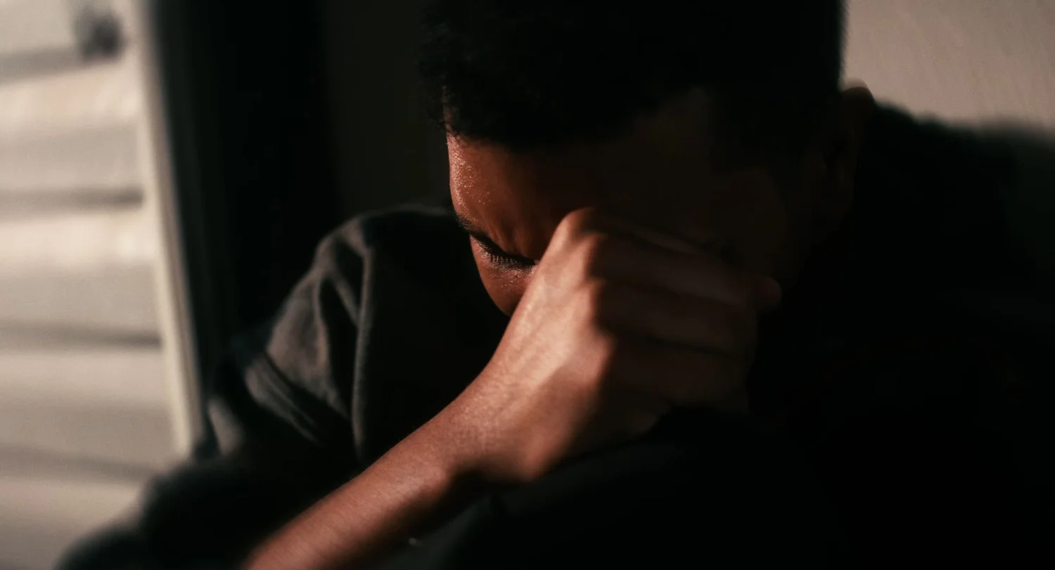 Still from INERTIA: a close view of a man in shadow pressing his fist to his forehead with his eyes closed, light from window blinds on the left.