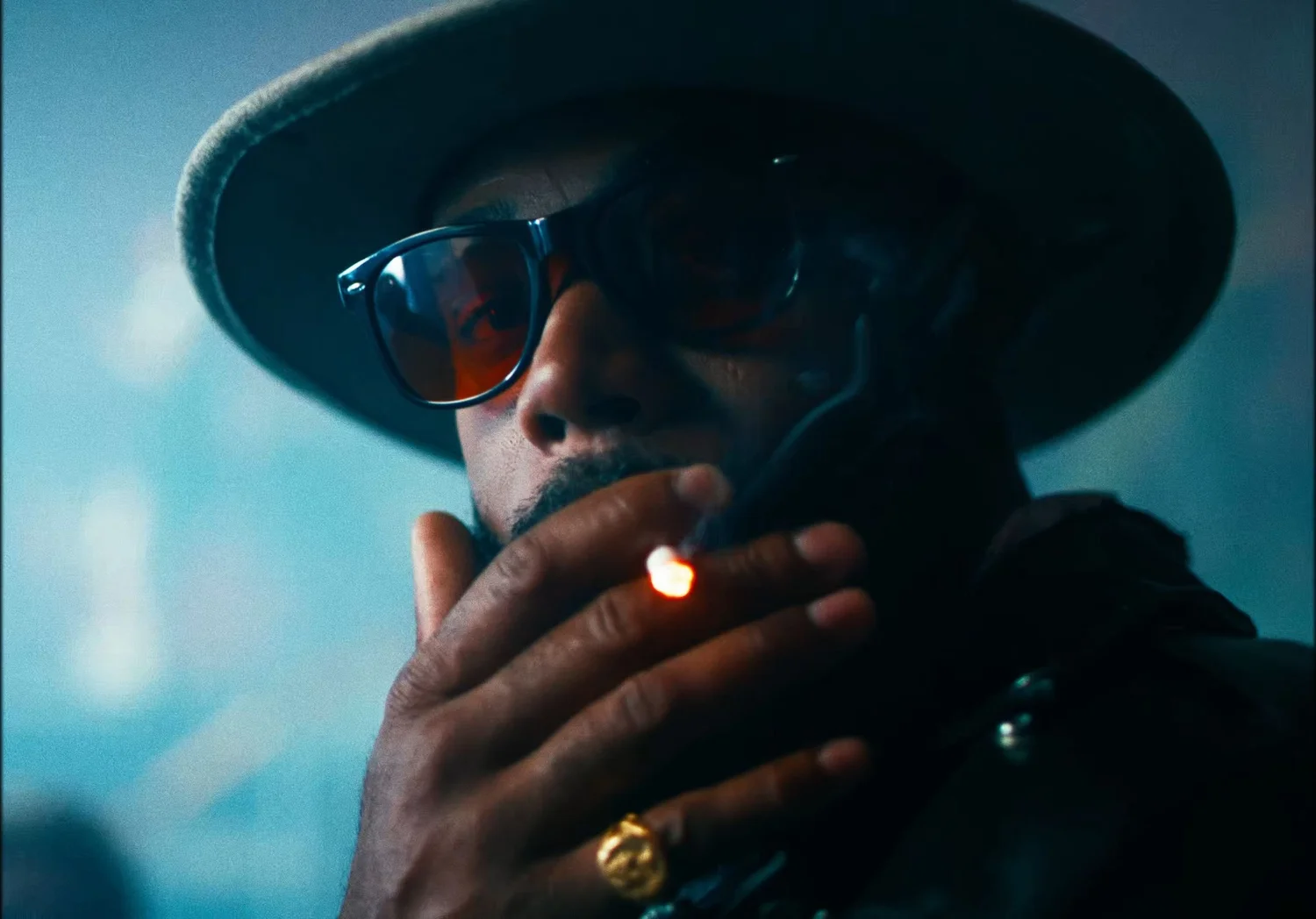 Still from INERTIA: a close view of a man in a wide brimmed hat and sunglasses holding a lit cigarette to his mouth, a gold ring on his finger.