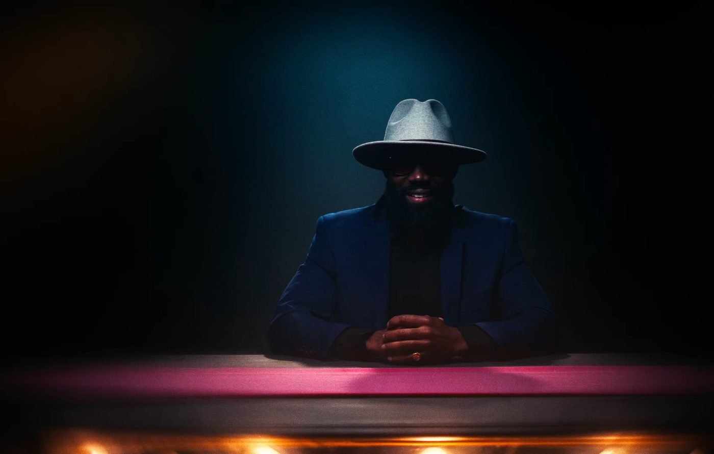 Still from INERTIA: a bearded man in a gray fedora, sunglasses and a blue jacket sits with his hands folded behind a desk with a pink stripe and a row of lit bulbs.