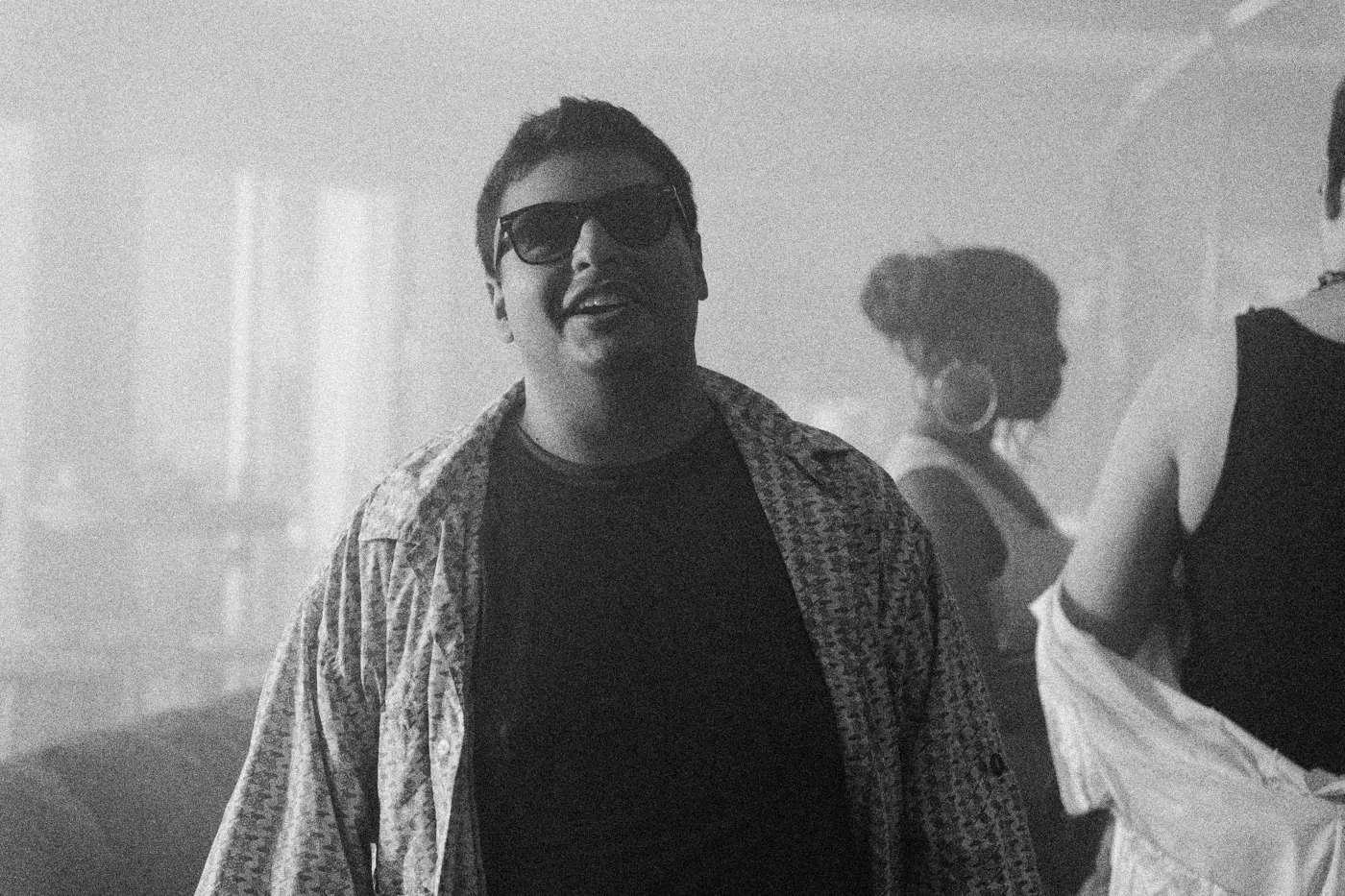 Behind the scenes on INERTIA: a man in sunglasses and an open patterned shirt grins in a hazy party room.