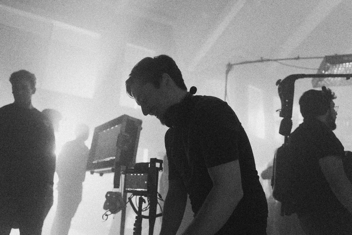 Behind the scenes on INERTIA: a crew member in red light leans over a monitor cart, other crew in haze behind.