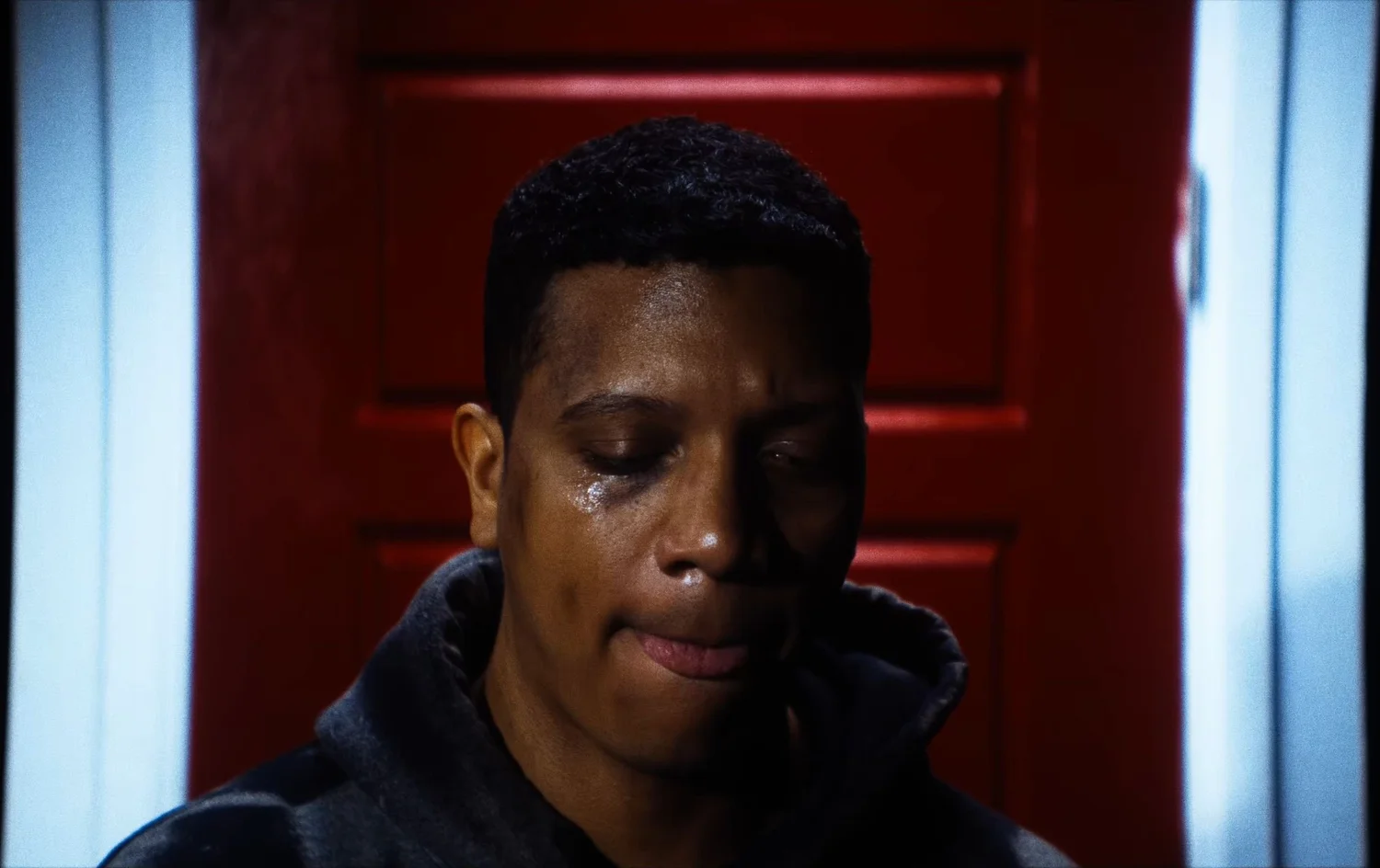 Still from INERTIA: a man in a dark hooded sweatshirt with a tear on his cheek stands before a red door framed in white.