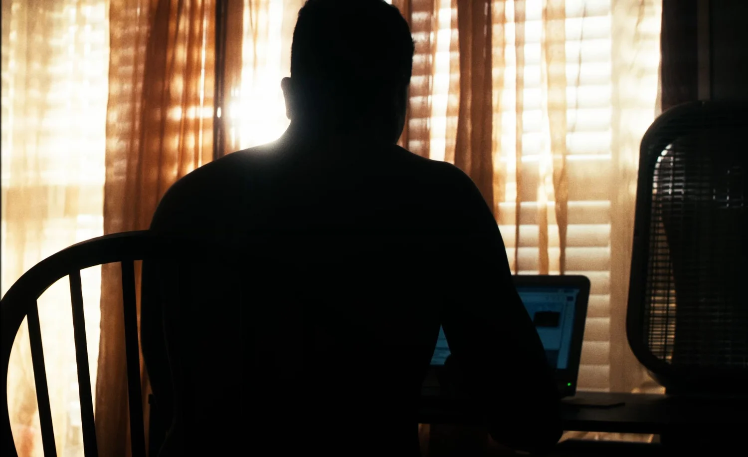 Still from INERTIA: a man in silhouette sits at a desk facing an open laptop, sunlight glowing through blinds and orange curtains, a box fan at the right.