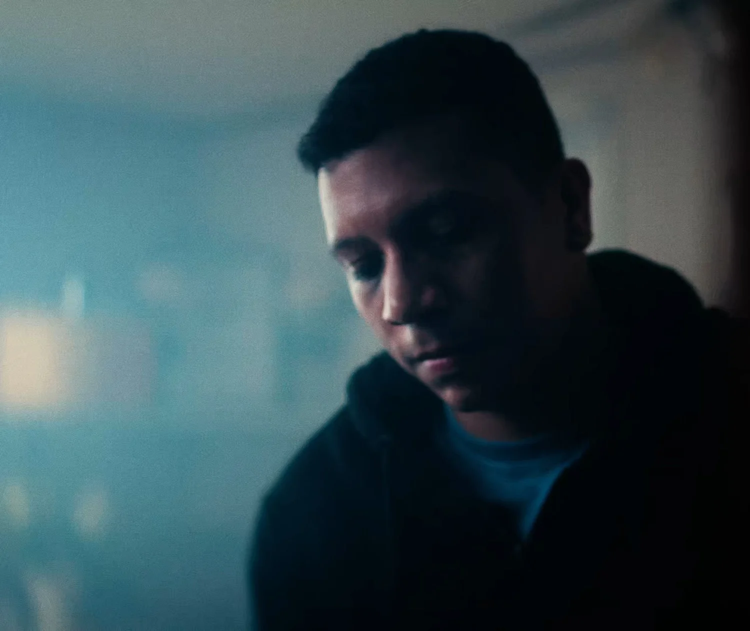Still from INERTIA: a man in a dark hooded sweatshirt looks down, soft blue haze filling the left side of the frame.