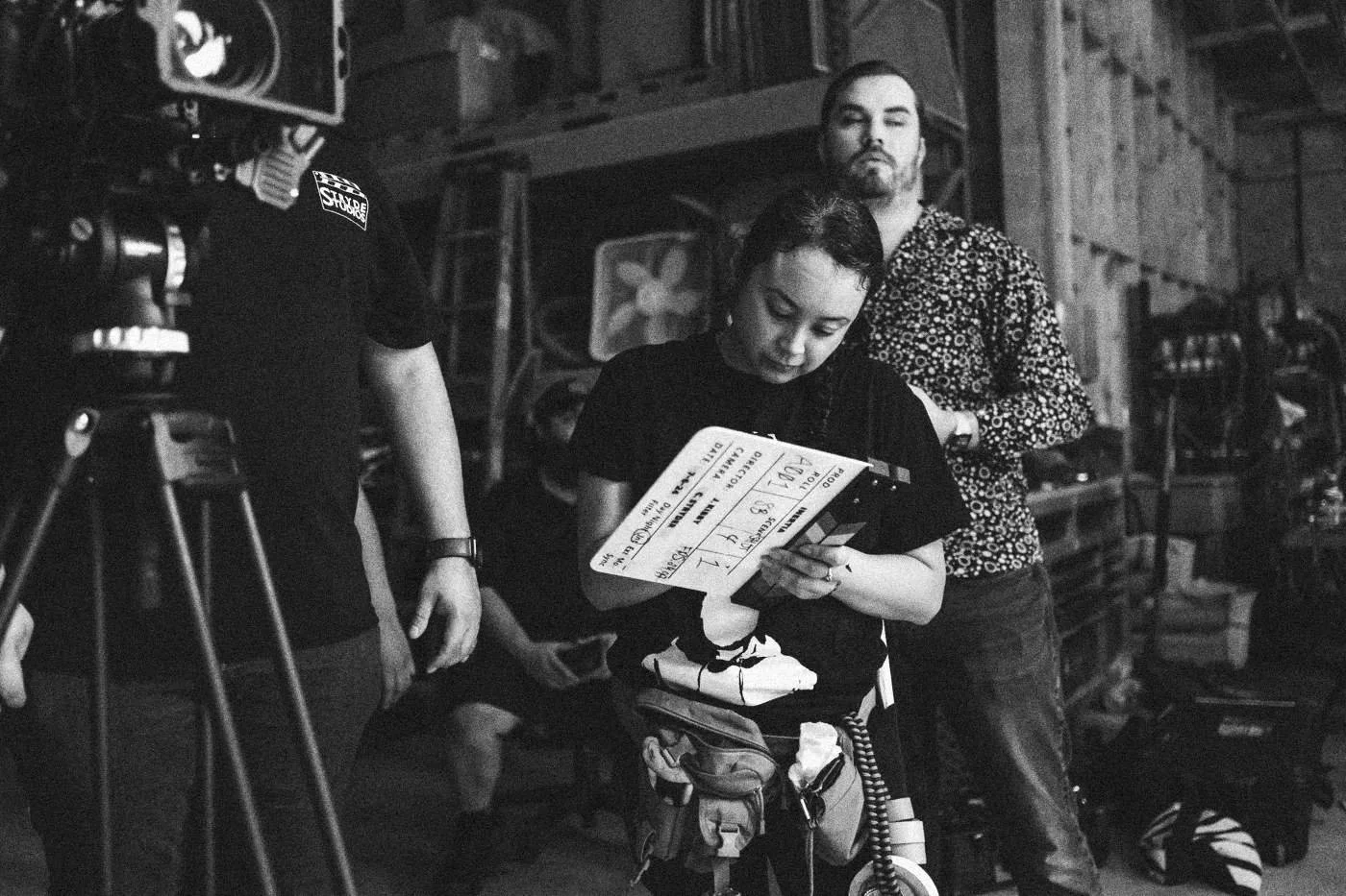 Behind the scenes on INERTIA: a crew member reads the slate before a take in a warehouse, crew around her.