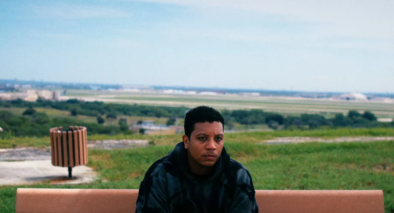 Still from INERTIA: a man in a dark hooded jacket sits on a bench on a grassy hilltop, flat land and a distant city under an overcast sky.