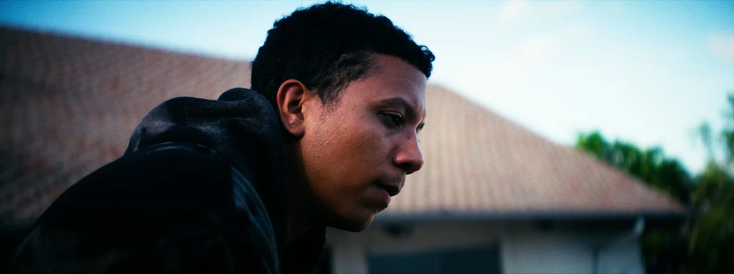 Still from INERTIA: a young man in a dark hooded sweatshirt leans forward in profile, a tiled roof and blue sky behind him.