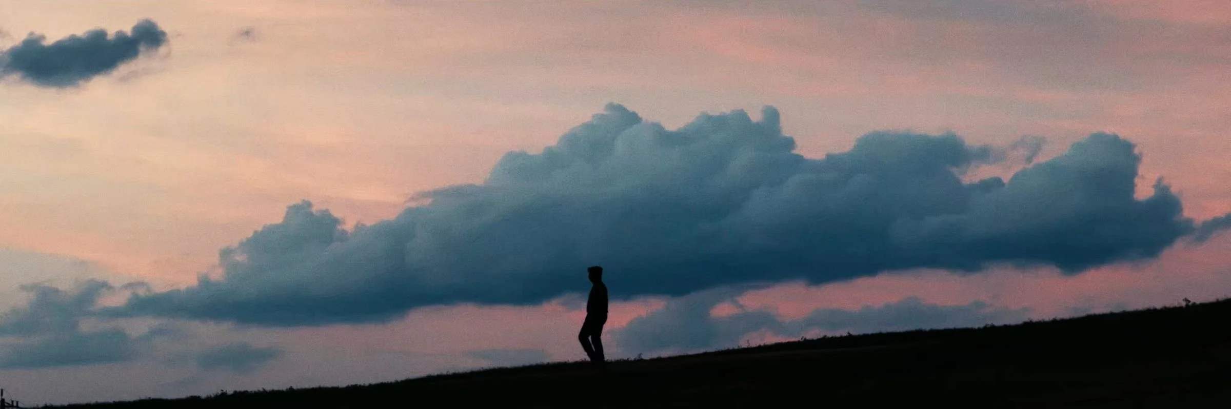 Still from INERTIA: a lone figure in silhouette walks along a hilltop under a long cloud at dusk.