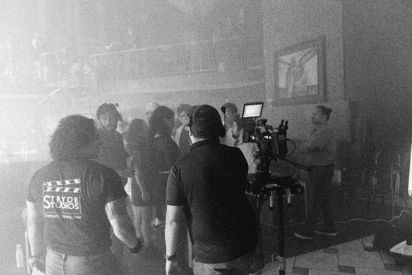 Behind the scenes on INERTIA: crew members in slate branded shirts watch a camera rig film the party scene through haze.
