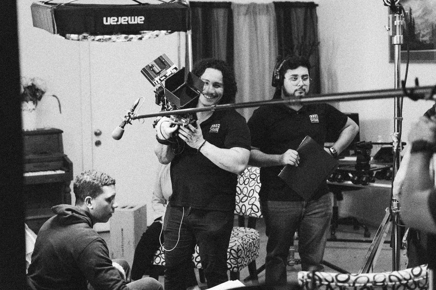 Behind the scenes on INERTIA: a smiling camera operator and a boom operator work on the living room set beside a crew member with a blue folder.