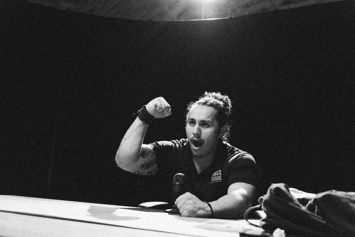 Behind the scenes on INERTIA: an actor raises a fist at a pink table under a single overhead light.