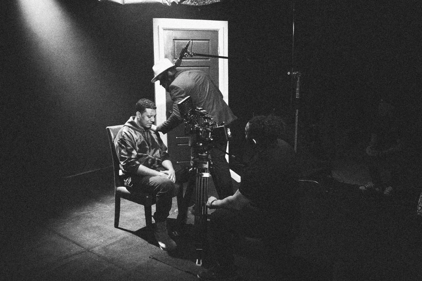 Behind the scenes on INERTIA: a man in a hat and blue suit leans over a seated actor beside a red door while a camera rolls.