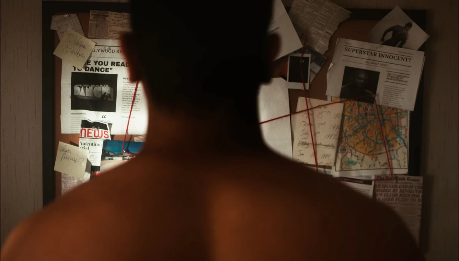 Still from INERTIA: a shirtless man seen from behind faces a corkboard covered in newspaper clippings, notes, photos and a map linked with red string.