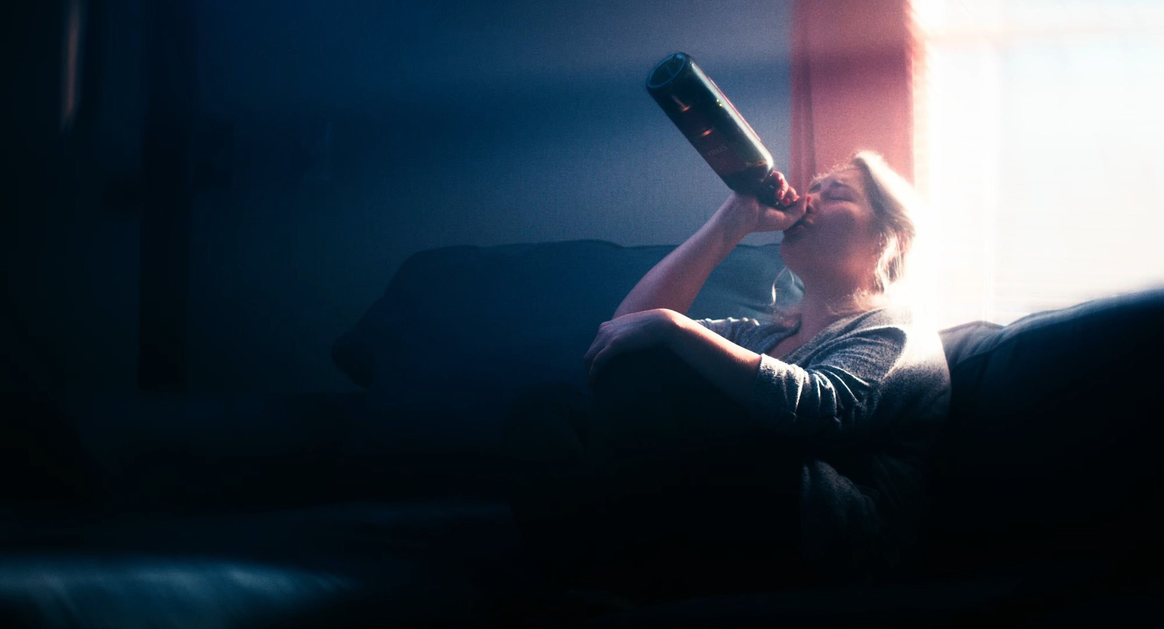 Still from INERTIA: a woman on a dark couch drinks straight from a wine bottle in the light of a bright window.