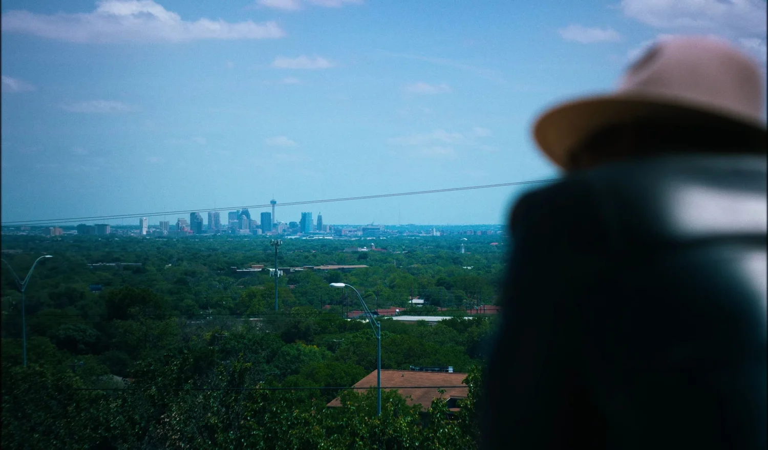 Still from INERTIA: a blurred figure in a wide brimmed hat stands in the right foreground facing a distant city skyline with a tall observation tower, green treetops and power lines between them.