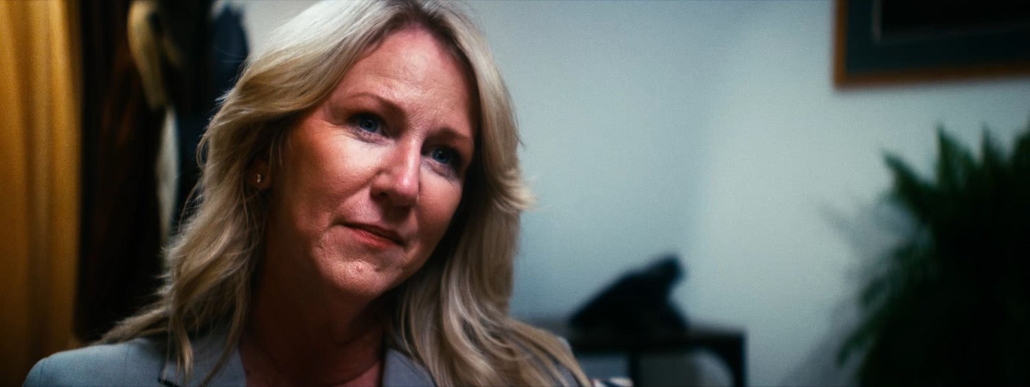 Still from INERTIA: a woman with long blonde hair and a gray blazer tilts her head in a room with a plant and a framed picture behind her.