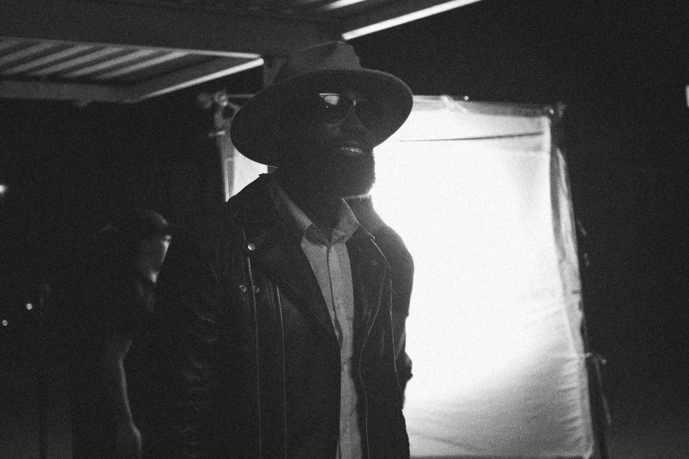 Behind the scenes on INERTIA: an actor in a wide brimmed hat and sunglasses smiles against a glowing diffusion frame at night.