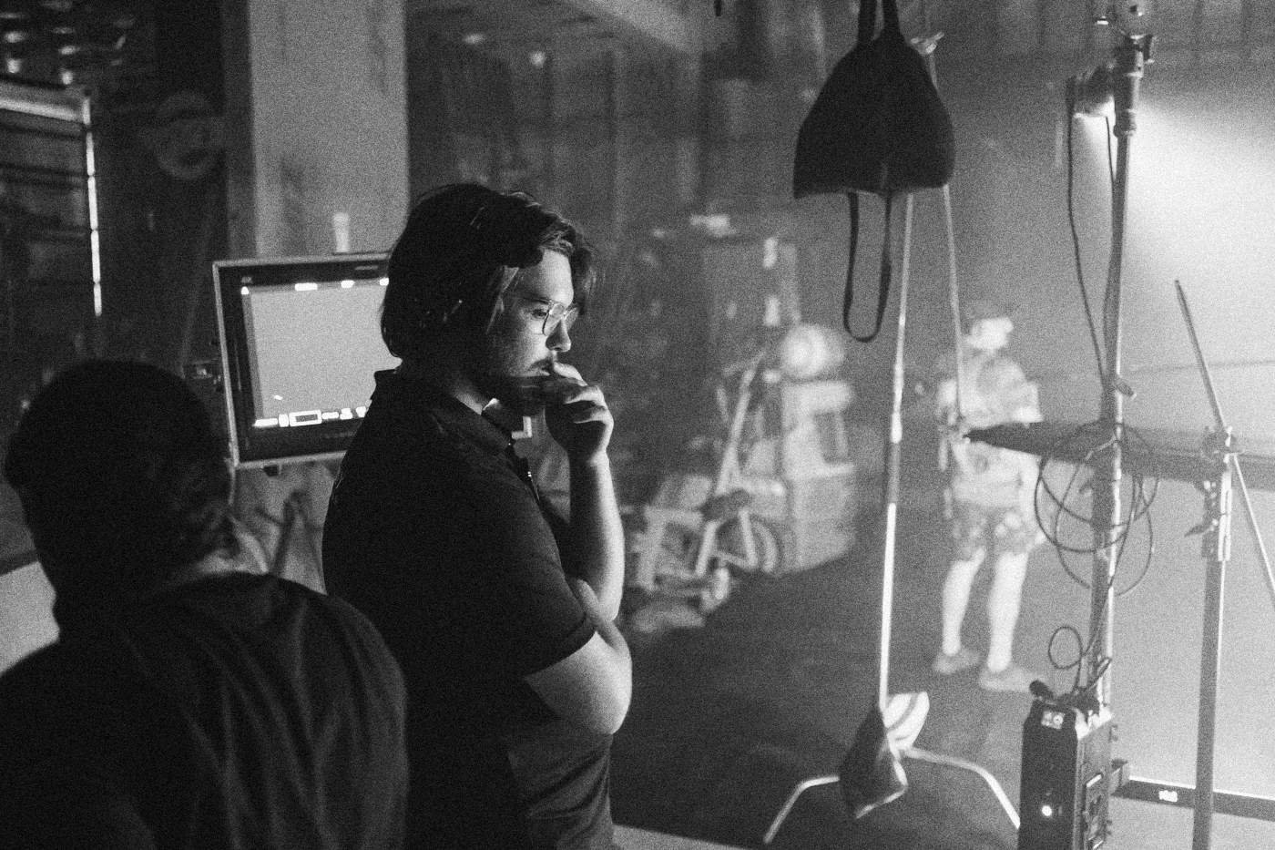 Behind the scenes on INERTIA: a crew member in a headset rests his hand on his chin beside a monitor in a workshop.