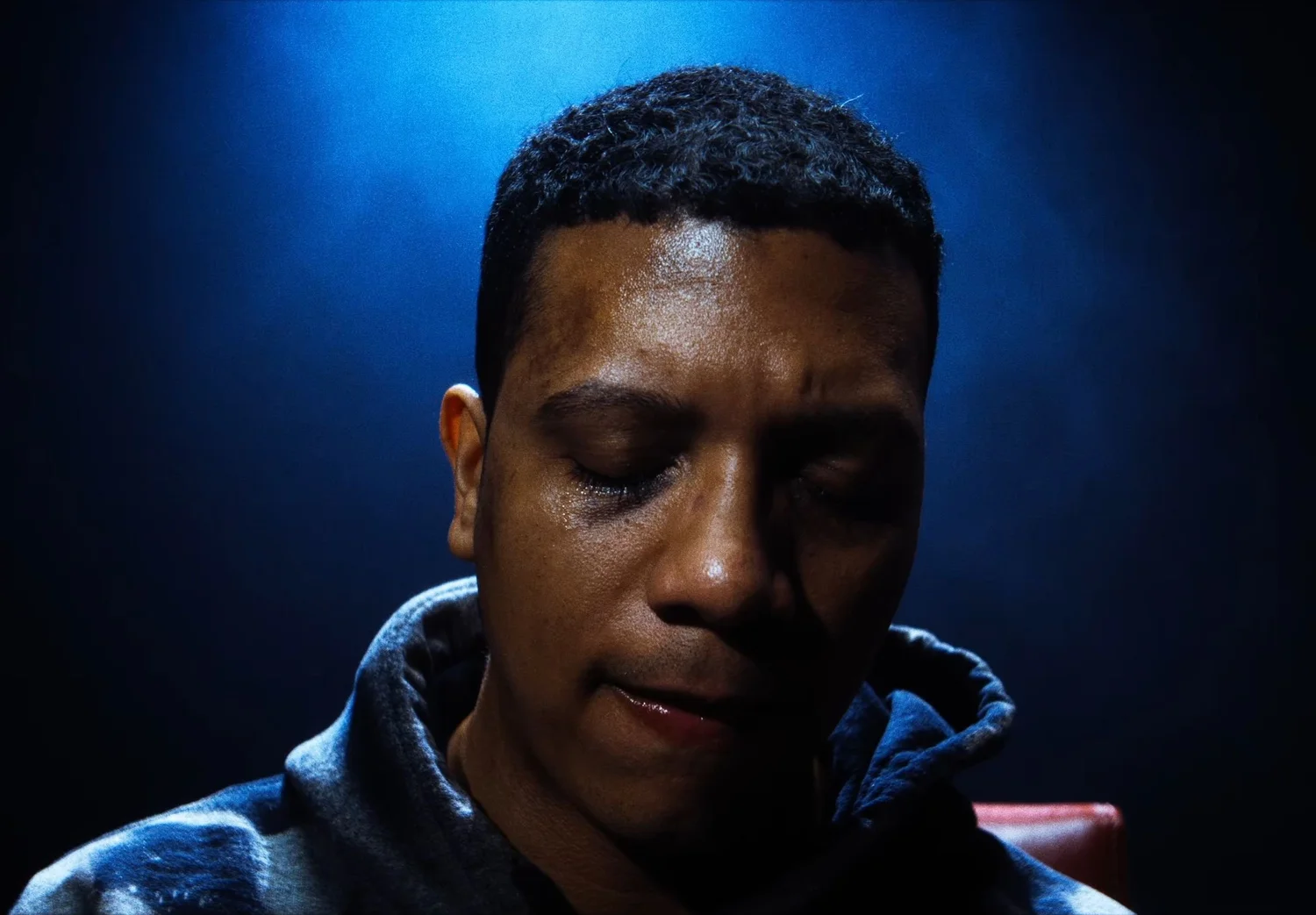 Still from INERTIA: a close view of a young man in a hooded sweatshirt with his eyes closed under blue light.