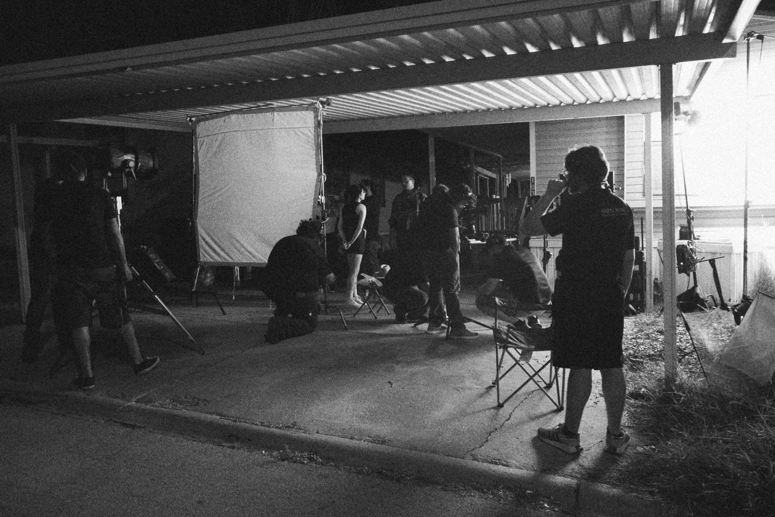 Behind the scenes on INERTIA: at night a film crew works under a carport around a large white diffusion frame, with lights on stands and folding chairs spread across the driveway.