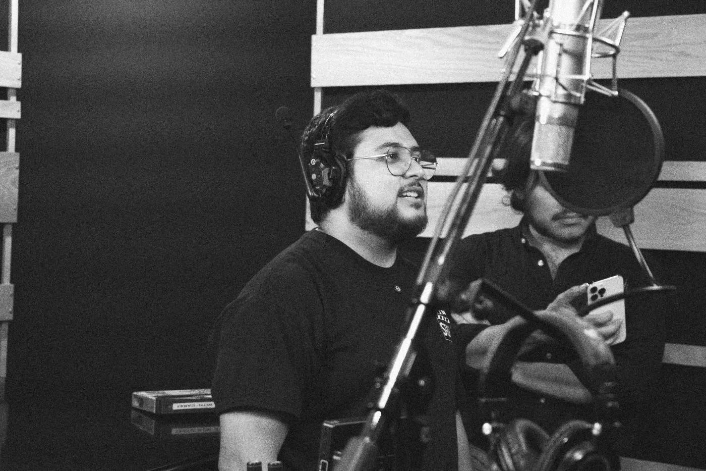 Behind the scenes on INERTIA: two crew members in headsets work beside a studio microphone, one checking a phone.