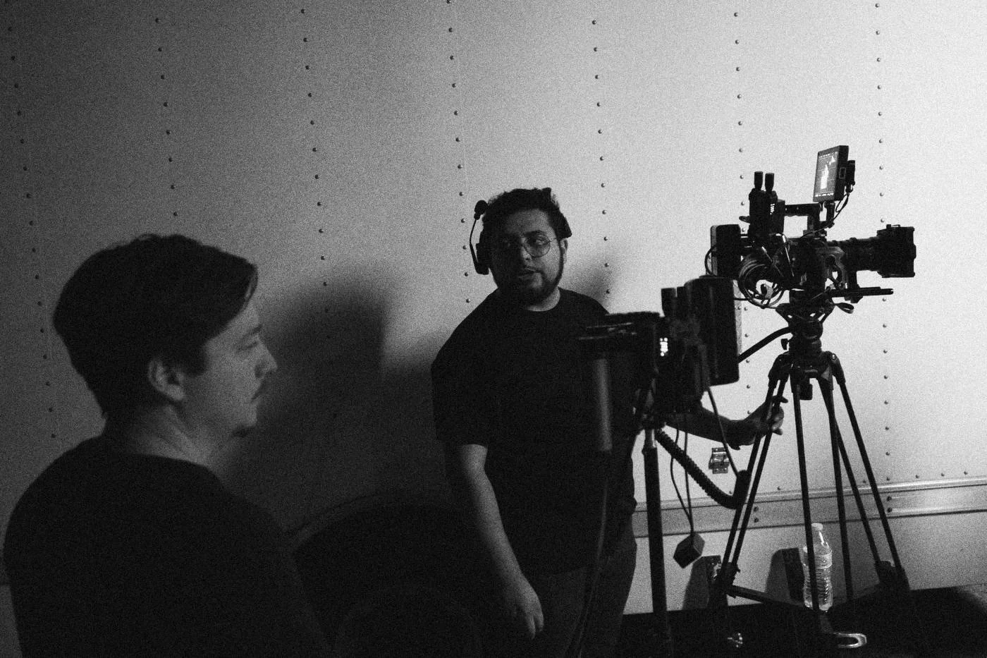 Behind the scenes on INERTIA: a camera operator on a tripod and a crew member work under hard light against a white wall.